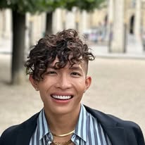 Beatus grinning, with curly dark hair, a dark blazer over a striped shirt, and a gold chain