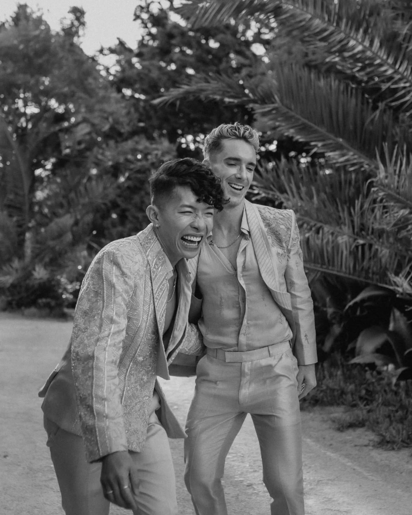 Black and white photo of Beatus and Mark in embellished suits, doubled over laughing under palm fronds