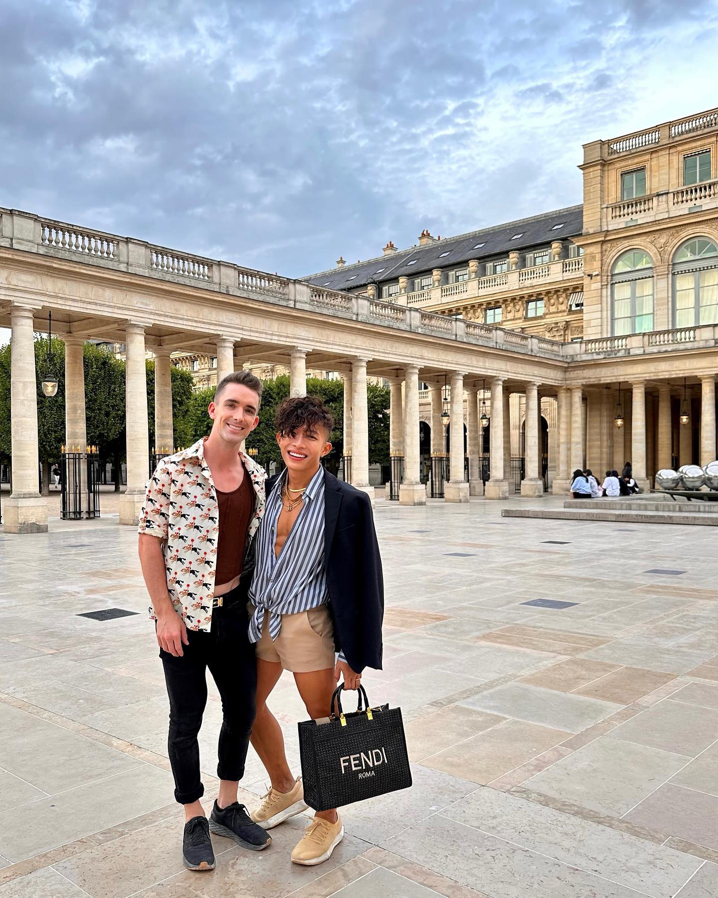 Mark and Beatus smiling in a grand stone colonnade courtyard, Beatus holding a black tote