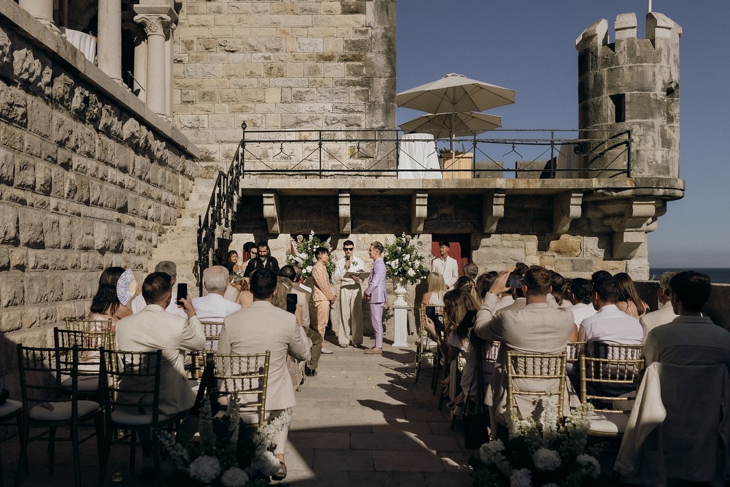 Wedding ceremony on a sunlit stone castle terrace by the sea, Beatus in peach and Mark in lavender facing each other before seated guests