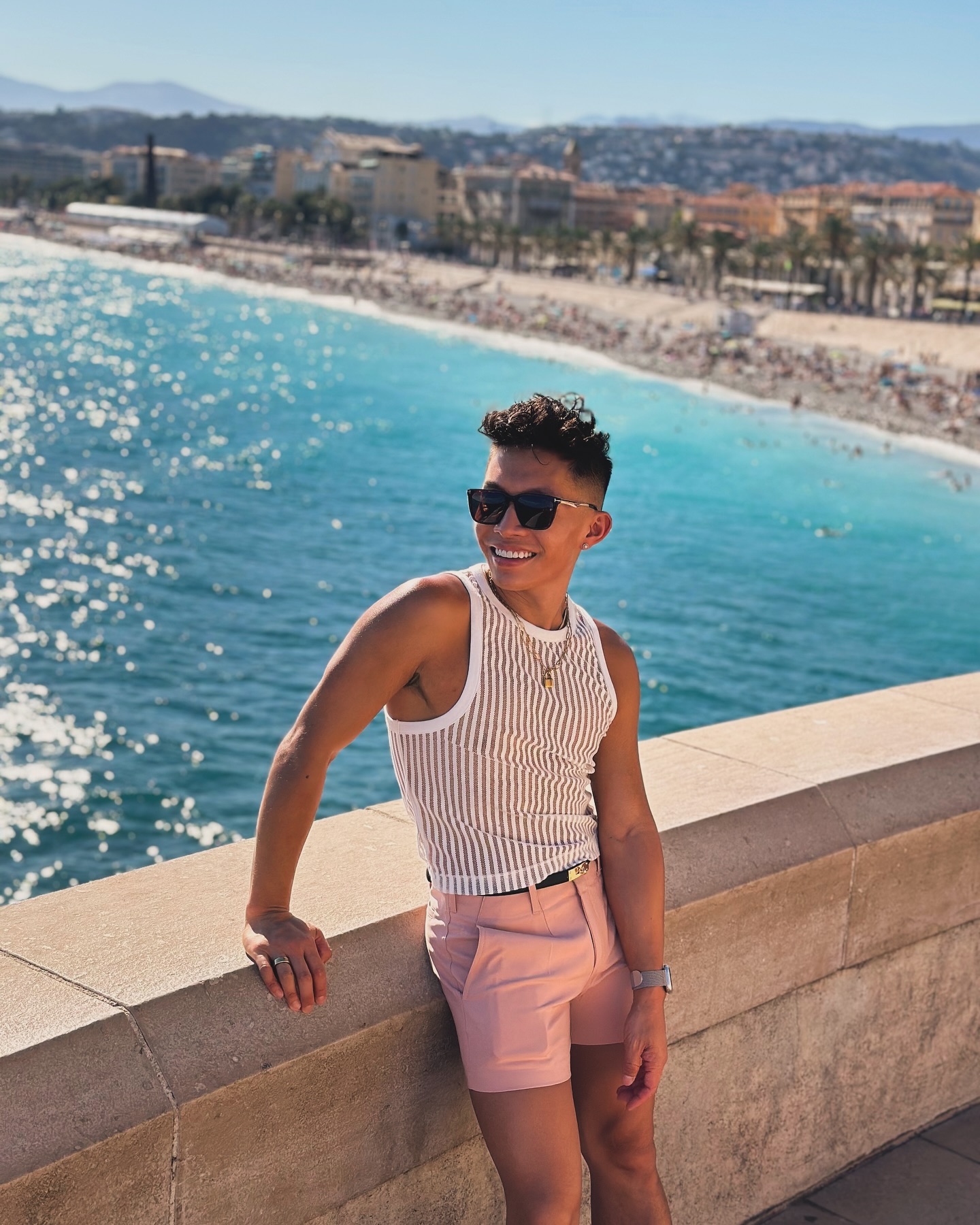Beatus in a sheer striped tank and pink shorts leaning on a stone wall above the turquoise water in Nice