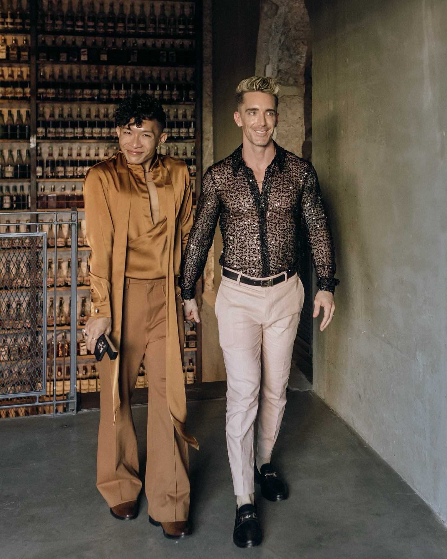 Beatus in a bronze satin suit and Mark in a sequined sheer shirt walking hand in hand past a wall of bottles