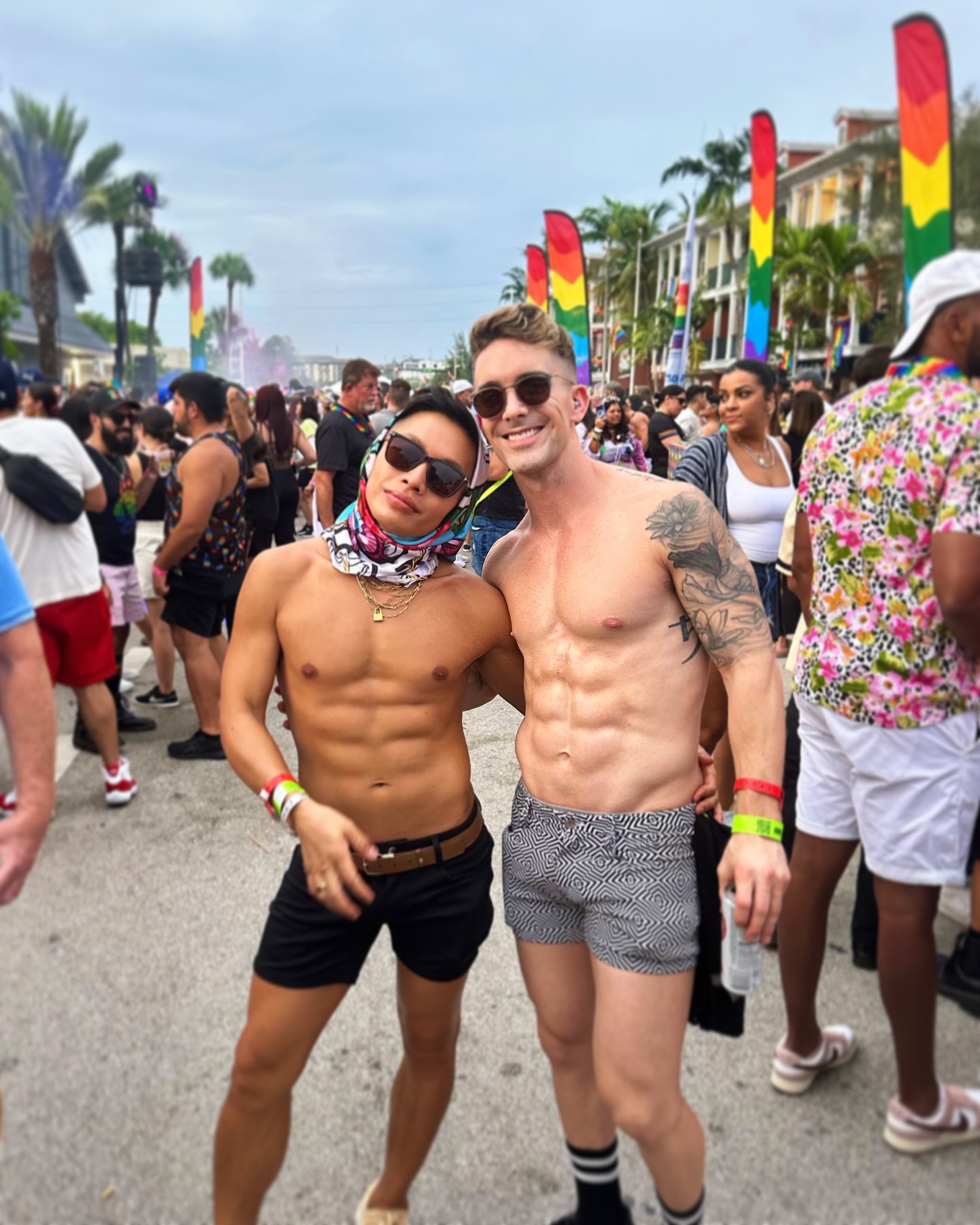 Beatus in a silk head scarf and Mark, both shirtless and grinning, in a crowded Pride street lined with rainbow flags and palms