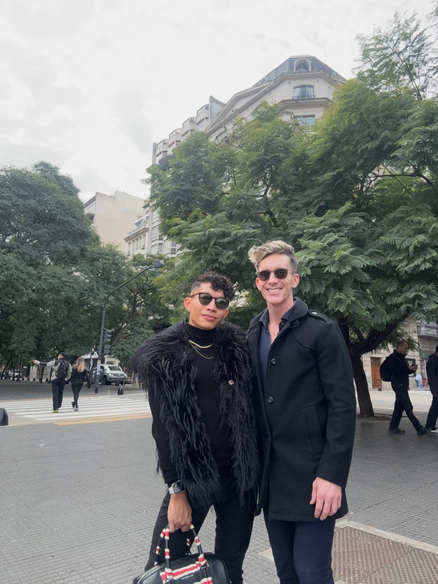 Beatus in a black feathered jacket and sunglasses with Mark in a black coat on a leafy Buenos Aires street